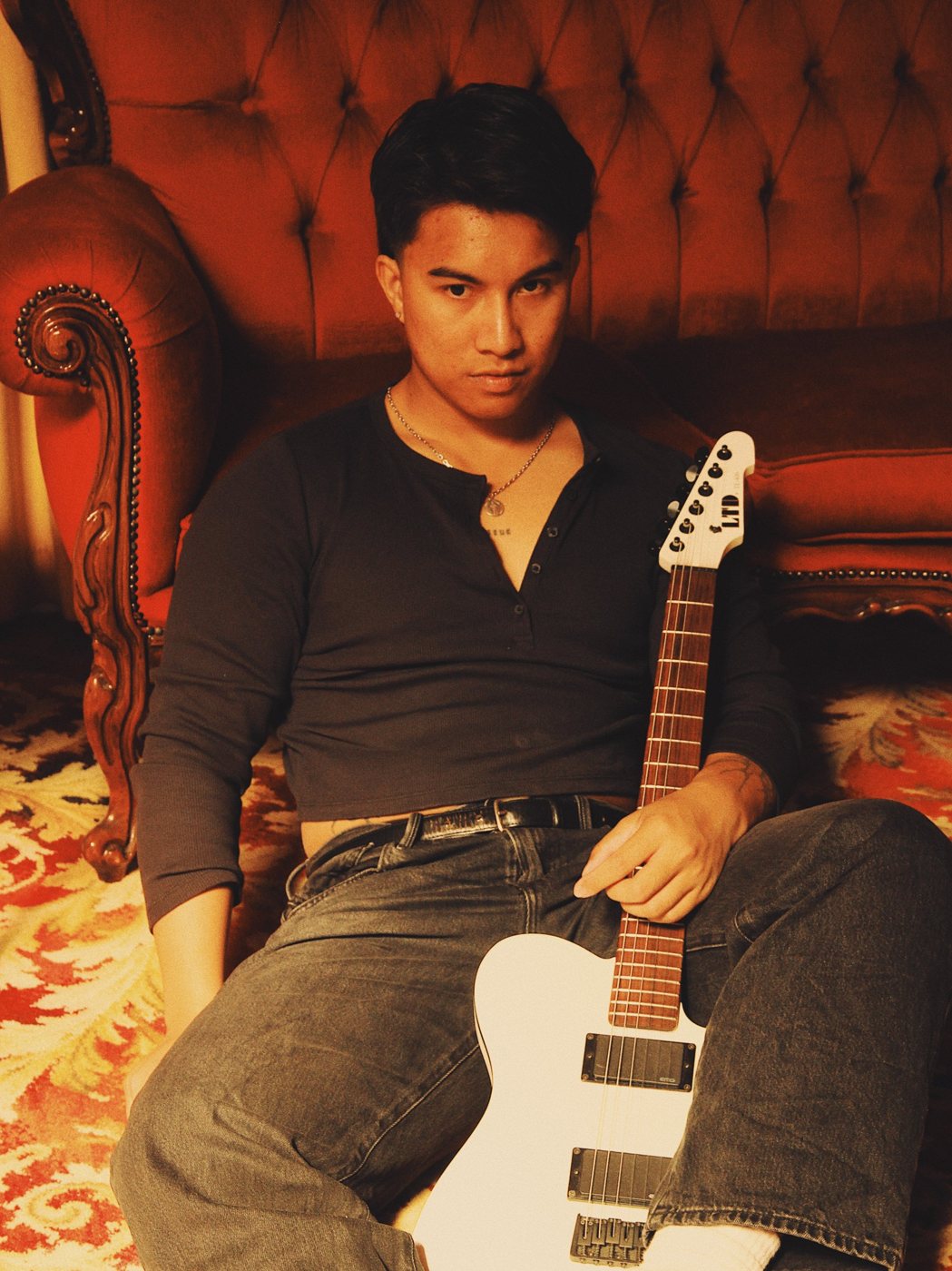 Aut sitting on the floor against a red velvet couch, black henley, white ESP LTD guitar, looking straight at the camera